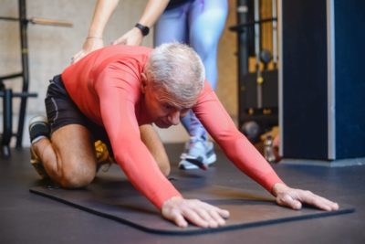 An older fit man stretches on a mat with a personal trainer nearby