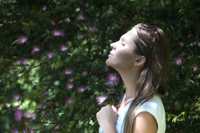 A woman stands with her eyes closed and pauses to take a deep breath in front of green plants with purple flowers